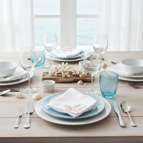 Two white napkins with embroidered blue and red coral designs on a gray surface with starfish and shells.